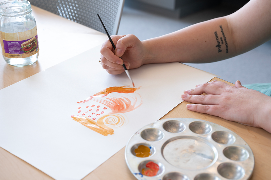 Morgan creates watercolor painting, orange paint on white paper with abstract swirls 